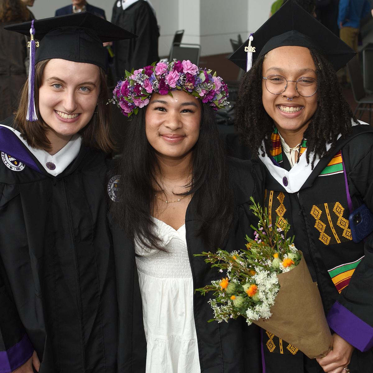 Three graduating students in robes, two in traditional caps and one wearing a beautiful flower crown, pose for a photo